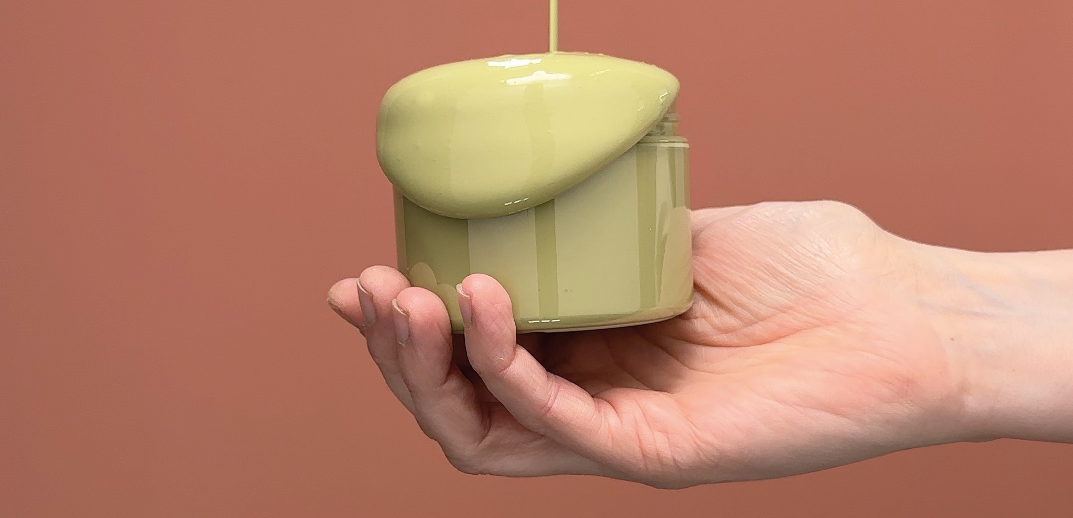 en hand h&aring;ller en burk pistagegr&ouml;n f&auml;rg mot en r&ouml;d bakgrund