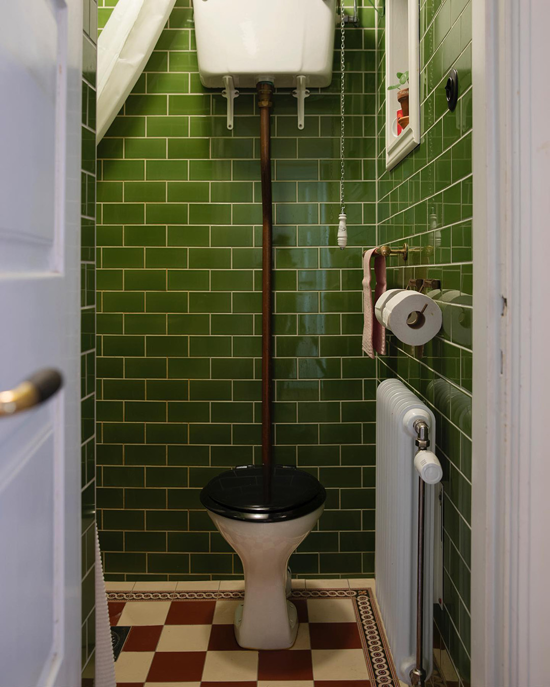 Badrum i sekelskiftesstil med v&auml;ggar av gr&ouml;nt kakel i half tileformat. Rutigt klinkergolv i r&ouml;tt och vitt med en m&ouml;nstrad b&aring;rd.