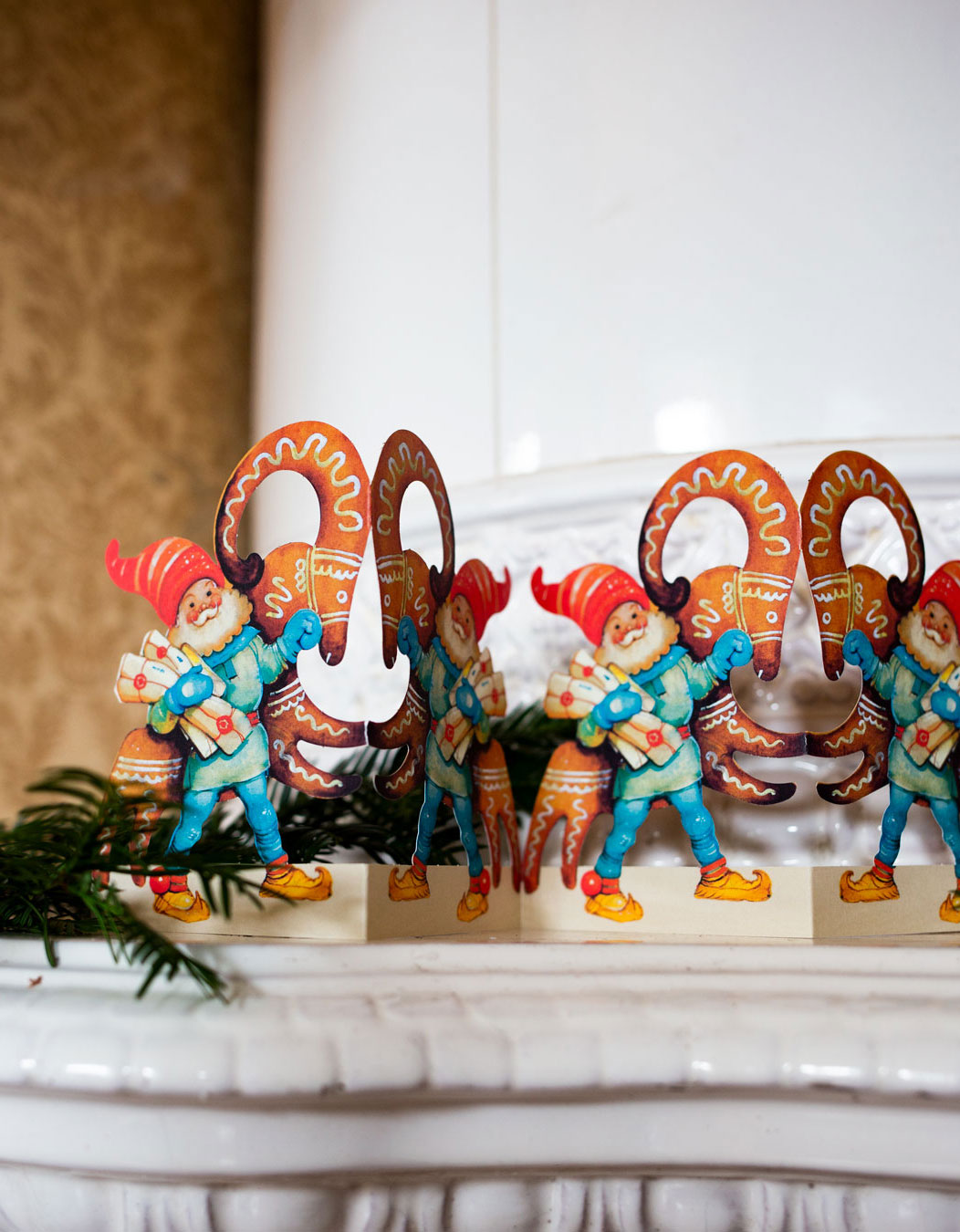 Pappersrad med motiv av tomte och julbock ovanp&aring; kakelugn