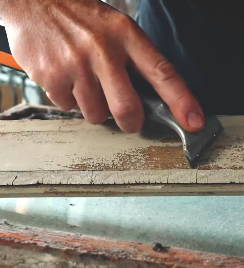 En hand skrapar bort gammal f&auml;rg fr&aring;n en f&ouml;nsterb&aring;ge.