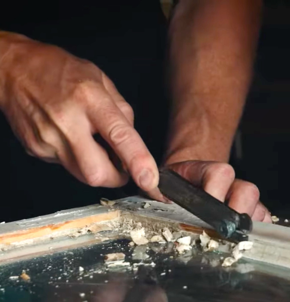 Tv&aring; h&auml;nder som skrapar en f&ouml;nsterb&aring;ge fr&aring;n gammalt kitt.