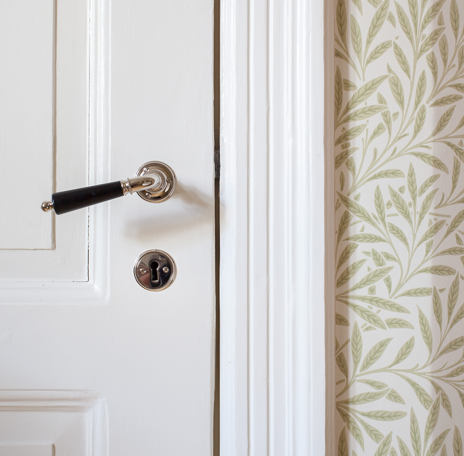 Ett f&ouml;rnicklat d&ouml;rrhandtag med svart tr&auml;grepp h&auml;nger ned&aring;t ist&auml;llet f&ouml;r att sitta rakt horisontellt. Vit spegeld&ouml;rr mot tapet med bladm&ouml;nster.